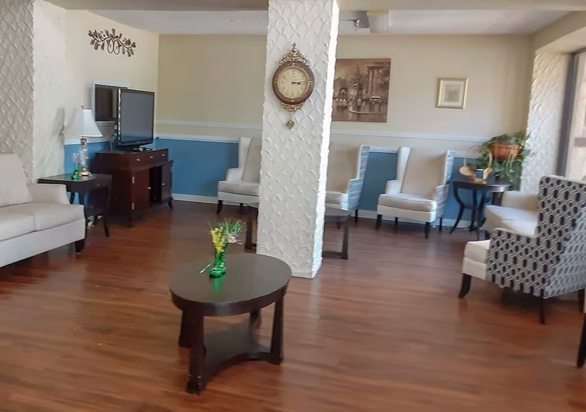 A cozy living room area with wooden flooring, several upholstered armchairs, a beige sofa, a dark wooden coffee table with a small green vase and flowers, a TV on a dark wooden stand, and decorative wall clock and artwork on textured white walls.
