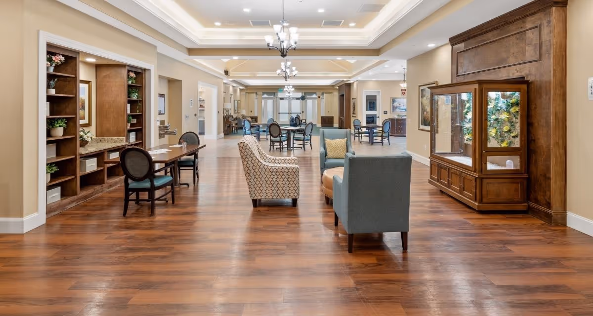 A spacious, well-lit common area in a senior living facility with wooden flooring, multiple seating arrangements including armchairs and tables with chairs, built-in wooden shelves with plants and decor, and a large wooden display cabinet on the right wall. The ceiling features recessed lighting and chandeliers.