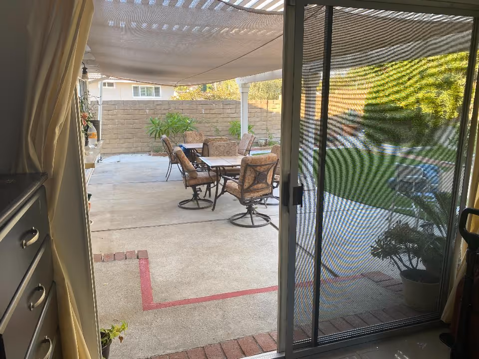 View through a sliding glass door with a mesh screen, looking out onto a covered patio area with a table and cushioned chairs. Beyond the patio is a small garden with green grass, plants, and a brick wall surrounding the yard.