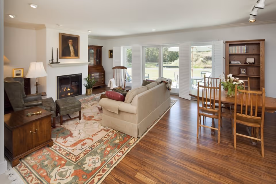 A cozy living room with a beige sofa, a green armchair with matching ottoman, a wooden rocking chair, and a wooden dining table with four chairs. The room features a fireplace with a painting above it, a patterned area rug, hardwood floors, and large windows with white shutters that open to a view of a green outdoor area.