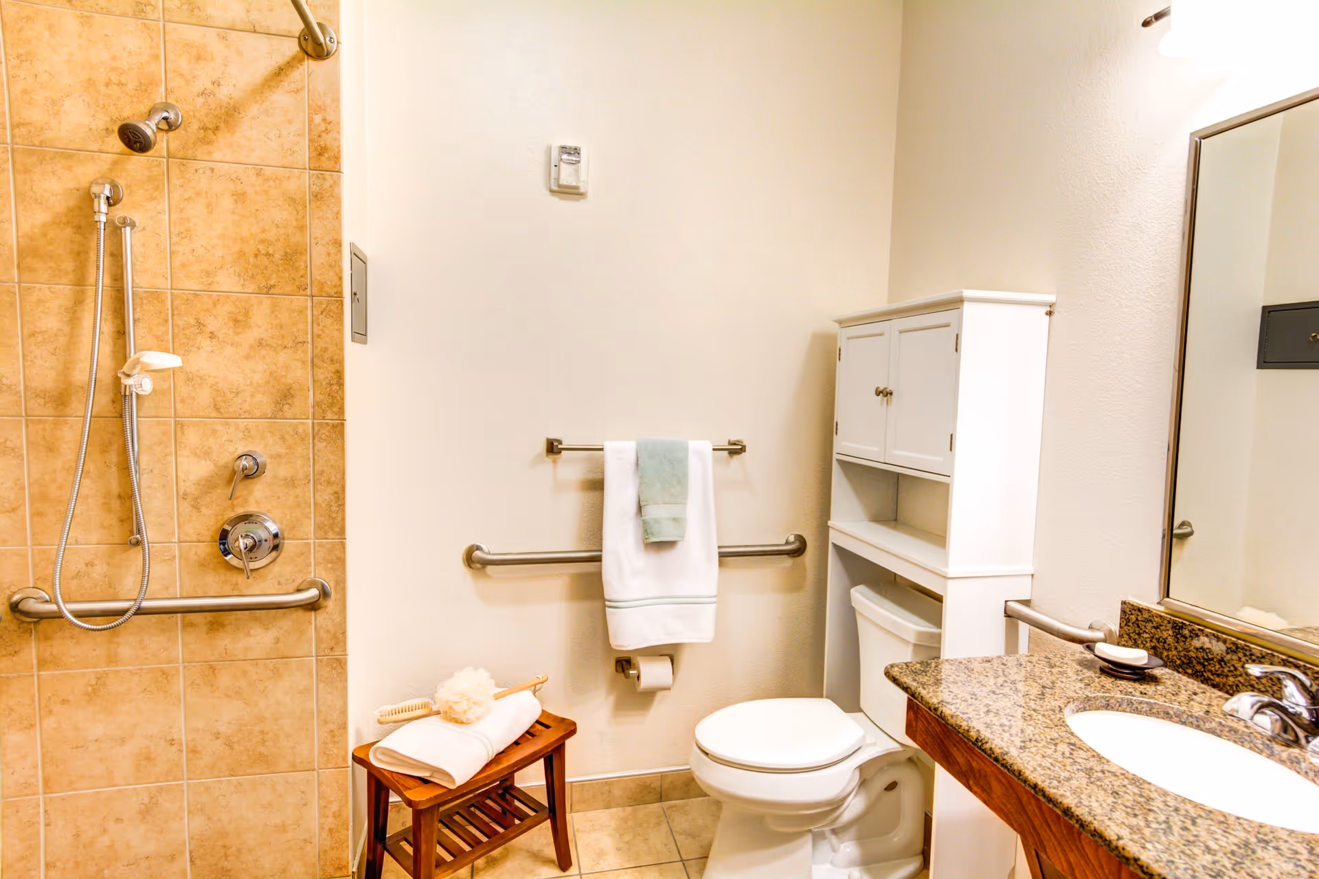 A clean and accessible bathroom featuring a tiled walk-in shower with grab bars and a handheld showerhead, a white toilet with a cabinet above it, a granite countertop with a sink, and a towel rack holding white and light blue towels. A small wooden stool with a folded towel and bath brush is also visible.