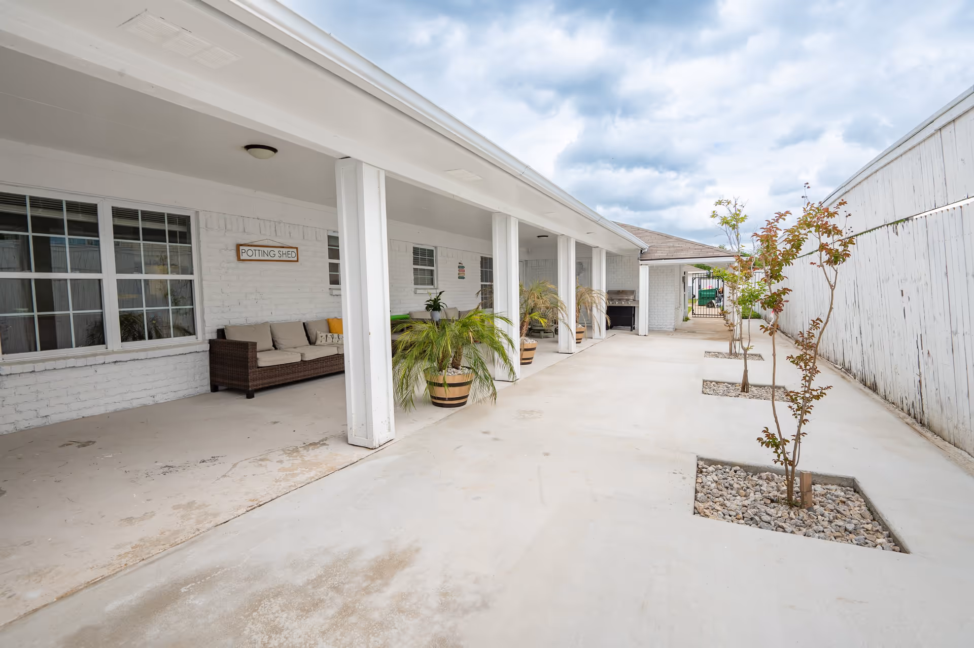 Outdoor covered patio area with white brick walls and columns, featuring a wicker sofa with beige cushions, potted plants, small trees planted in square rock beds along a concrete walkway, and a sign on the wall that reads 'POTTING SHED'.
