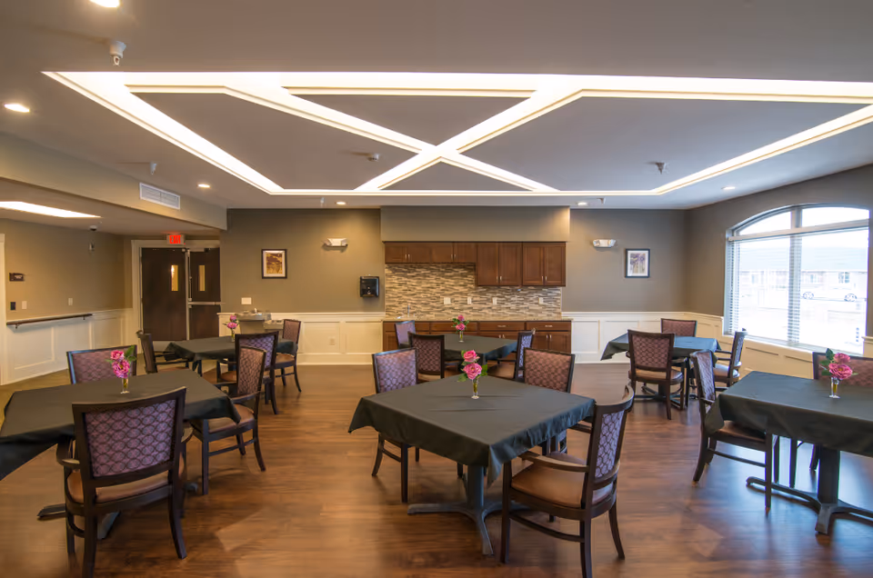 Bright dining room with multiple square tables covered in black tablecloths, purple-upholstered chairs, small flower centerpieces, and a kitchenette along the back wall.