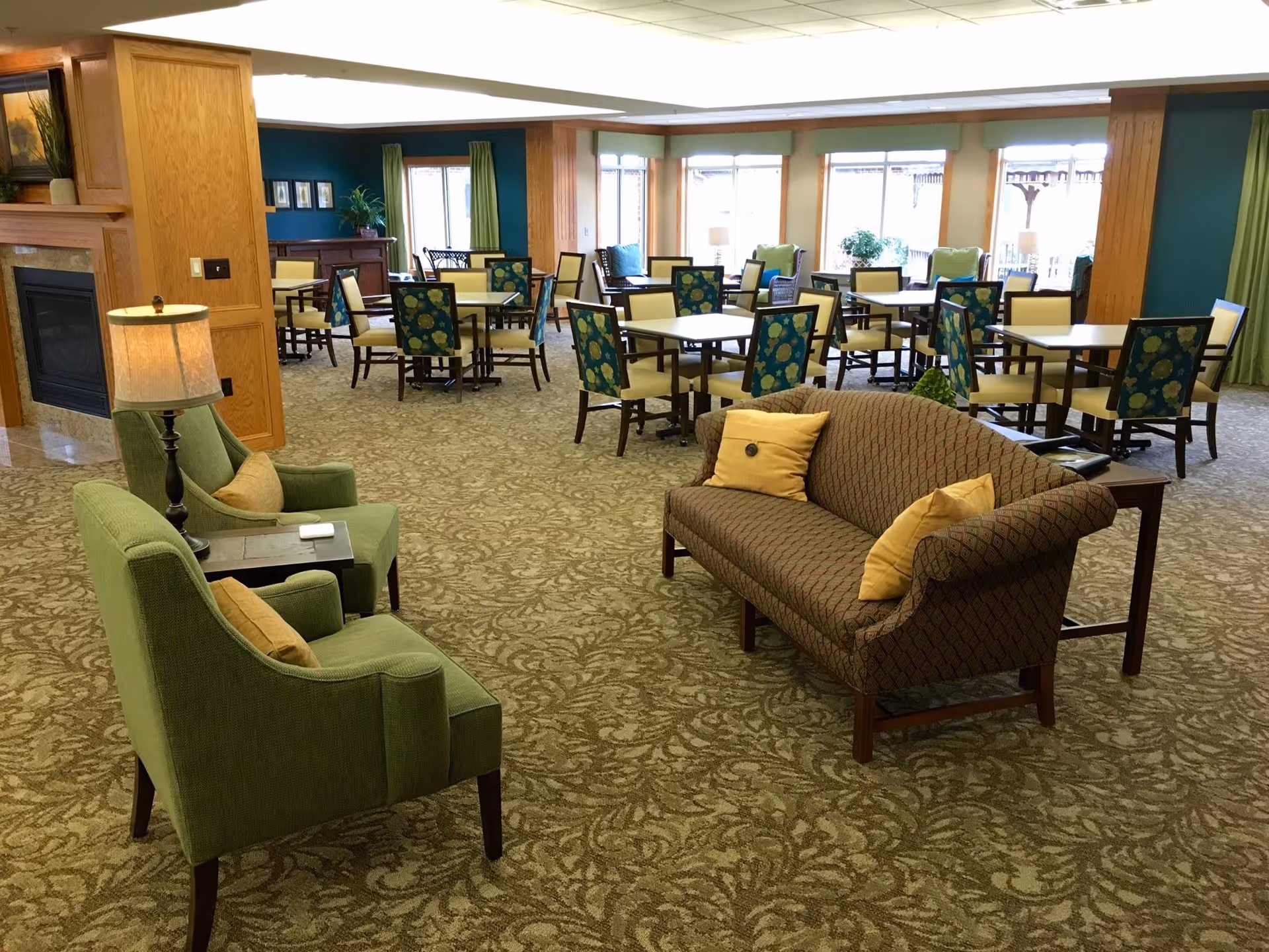 A spacious senior living common area with patterned carpet, several tables and chairs with floral upholstery, a brown patterned sofa with yellow pillows, and two green armchairs with a small table and lamp between them. Large windows in the background let in natural light.