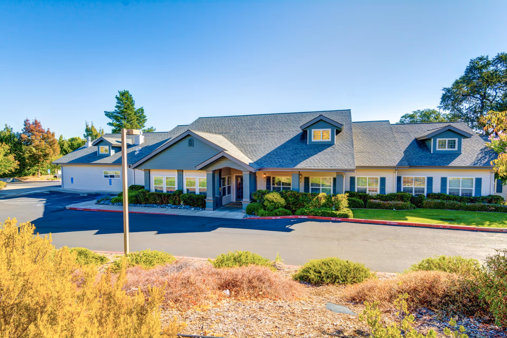 Exterior view of a single-story senior living facility building with a gray roof, multiple windows, and a covered entrance. The building is surrounded by well-maintained landscaping including bushes, trees, and dry plants in the foreground. The sky is clear and blue.