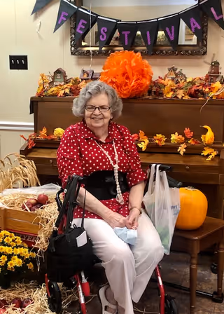 An elderly woman with gray hair and glasses, wearing a red polka dot blouse and white pants, is sitting on a walker in a decorated room. Behind her is a piano adorned with autumn-themed decorations including leaves, small houses, and a large orange pom-pom. A banner above the piano spells out 'FESTIVAL'. There is a pumpkin on a small table next to her and a basket with apples and hay on the floor nearby.