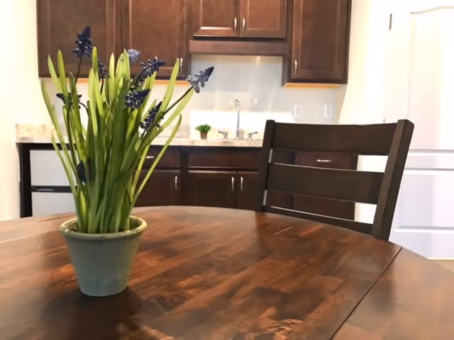 A wooden dining table with a small green potted plant with purple flowers on it. Behind the table is a dark wooden chair and a kitchen area with dark brown cabinets, a countertop, a sink, and a white refrigerator.