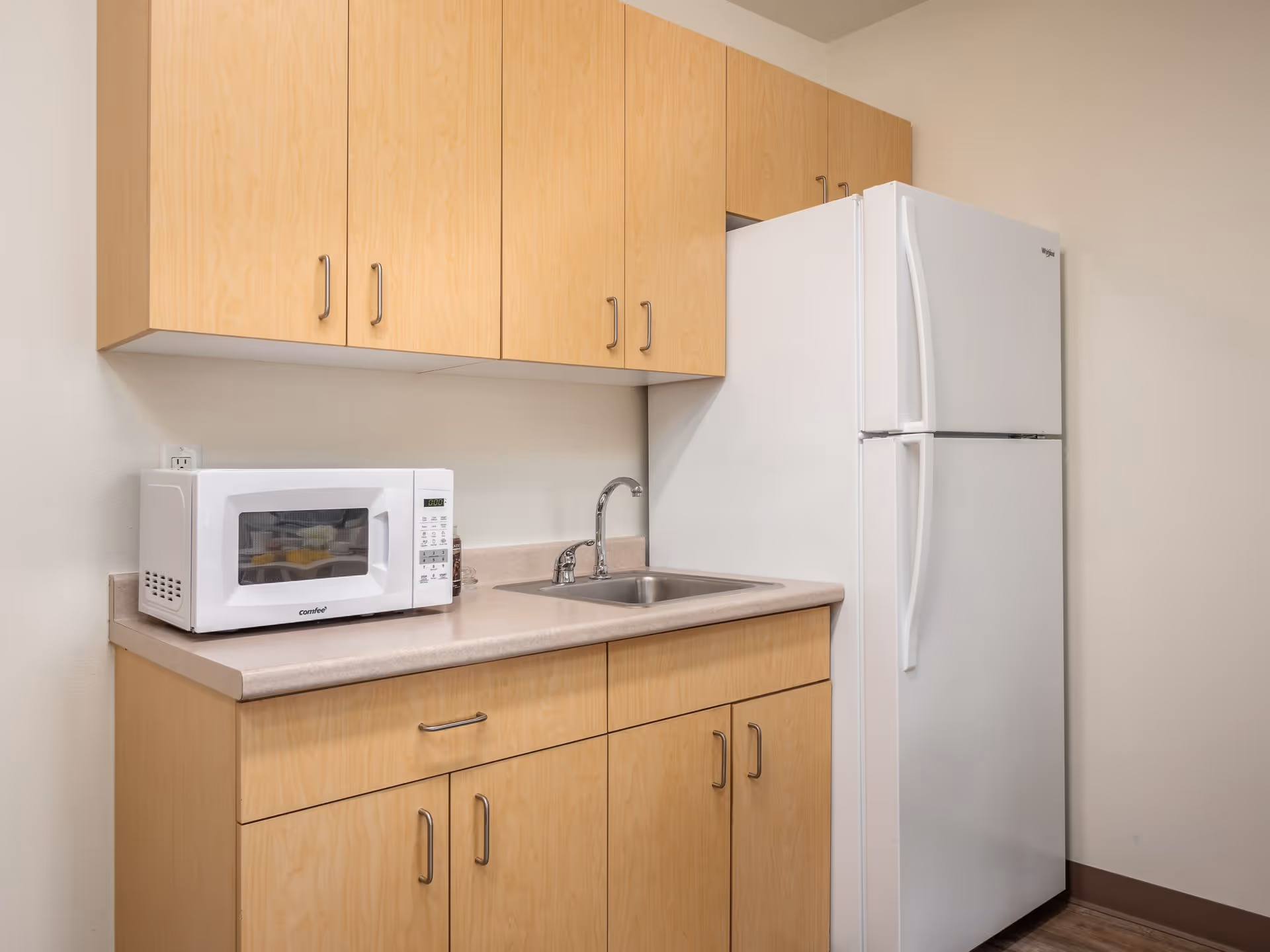 A small kitchen area with light wood cabinets, a white microwave on the countertop, a stainless steel sink with a faucet, and a white refrigerator against a beige wall.