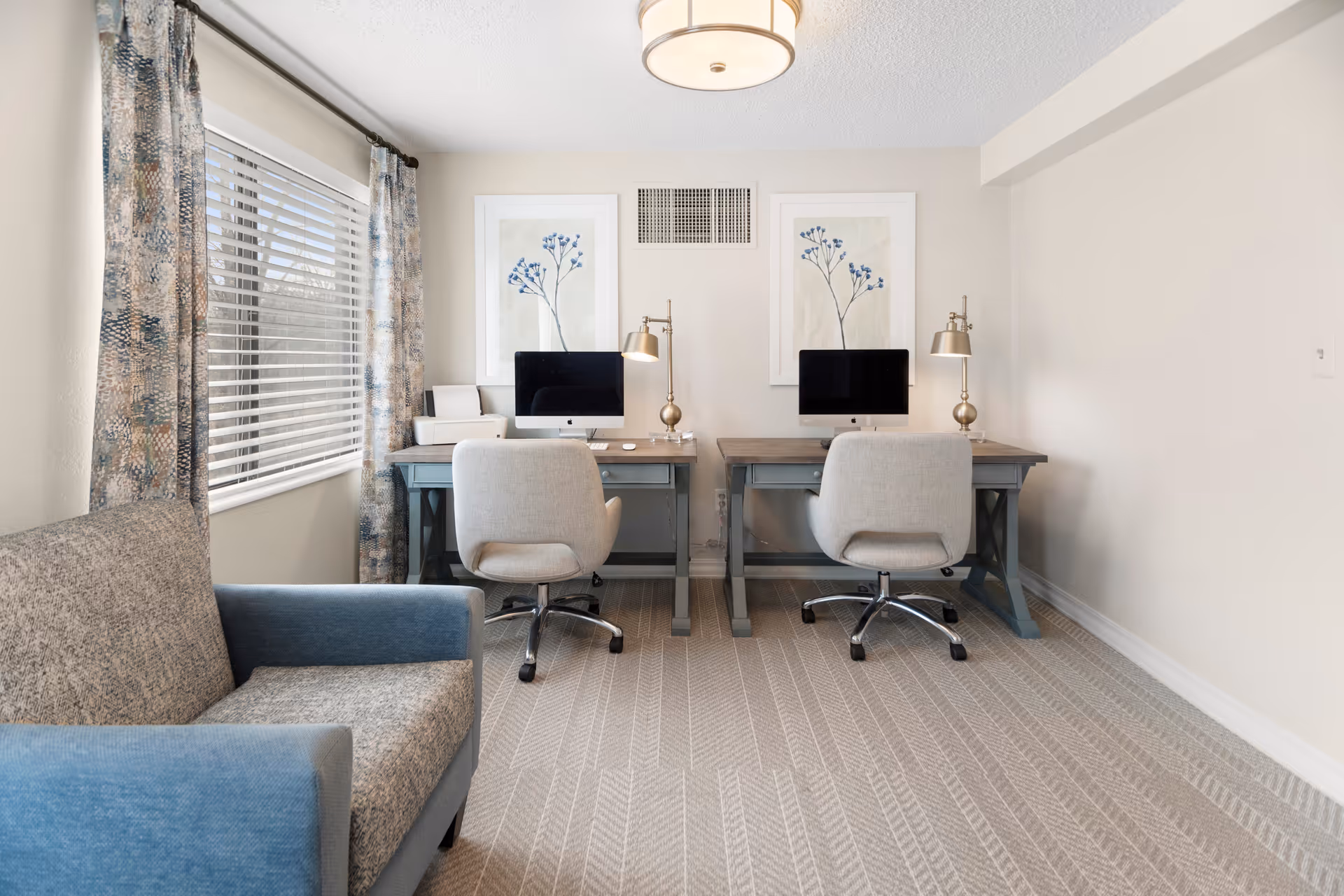 A bright room with two desks each holding a computer and a lamp, two cushioned office chairs, a window with patterned curtains, and a blue and gray upholstered sofa on the left side.