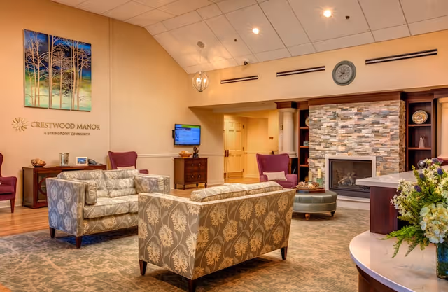 Lounge area with patterned sofas and armchairs facing a stone fireplace under a Crestwood Manor sign on the wall.