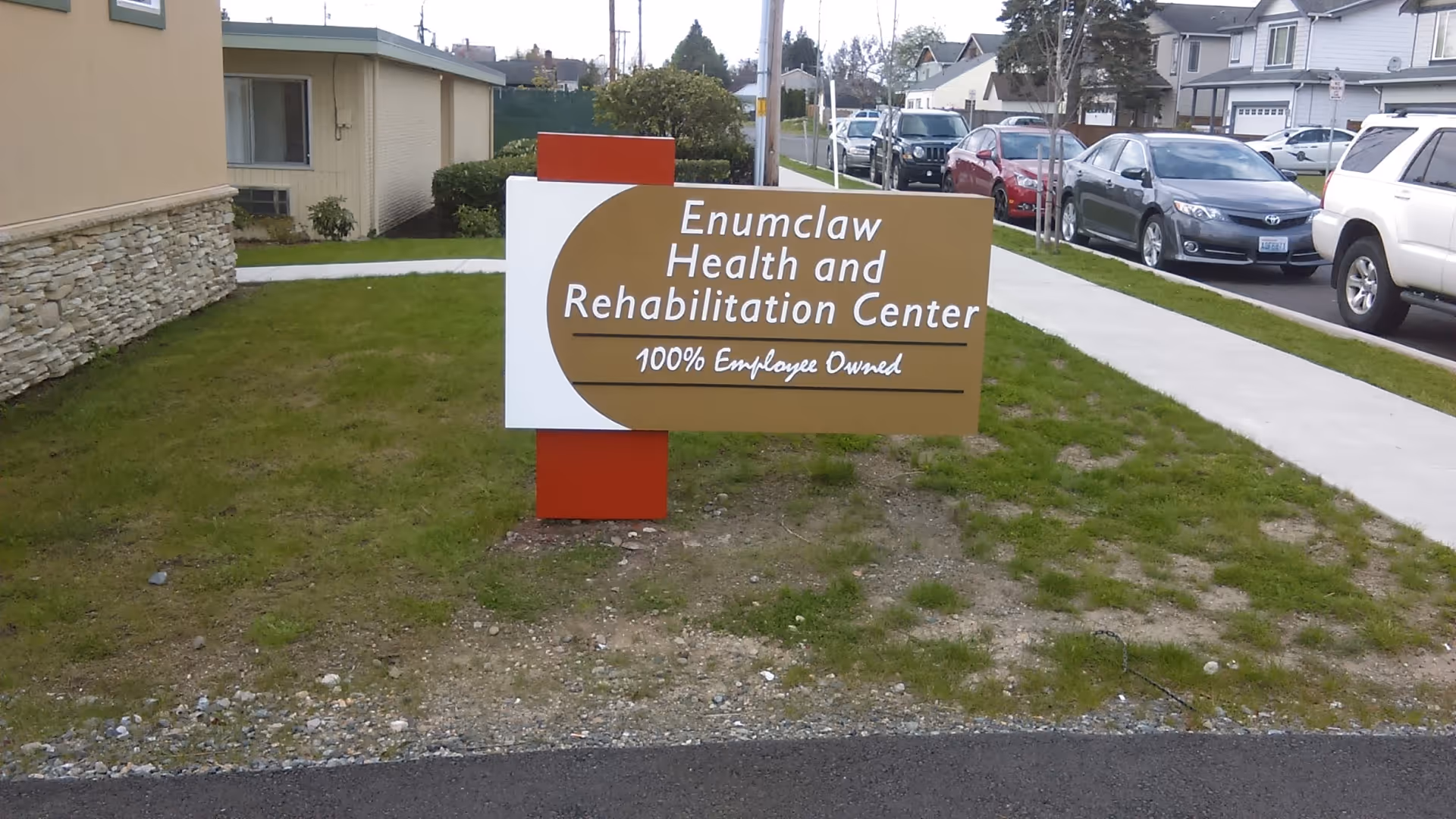 A lawn-mounted sign reading "Enumclaw Health and Rehabilitation Center 100% Employee Owned" in front of the facility with a sidewalk and parked cars.