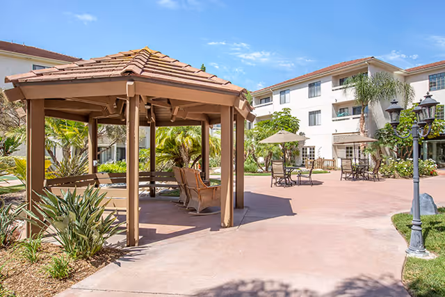 Outdoor courtyard area at Brookdale Irvine featuring a wooden gazebo with seating, patio tables with umbrellas, surrounded by greenery and a multi-story residential building under a clear blue sky.