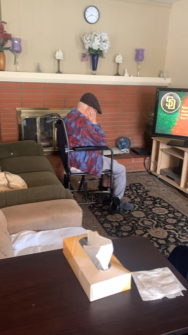 An elderly person sitting in a wheelchair facing a television in a living room. The room has a brick fireplace with decorative items on the mantle, a clock on the wall, a patterned rug, and a couch. A tissue box and some tissues are on a dark wooden table in the foreground.