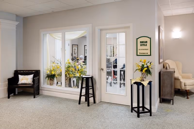 Interior view of a senior living facility hallway with a window and door leading to a hair salon named 'Shear Style' at Pinebrook. The hallway features a black bench with a decorative pillow, two black plant stands with yellow floral arrangements, a beige armchair, and soft lighting on the walls.
