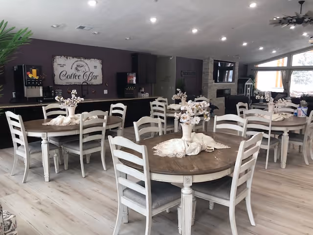 Bright communal dining room with round wooden tables and white chairs set with floral centerpieces and a coffee bar in the back.