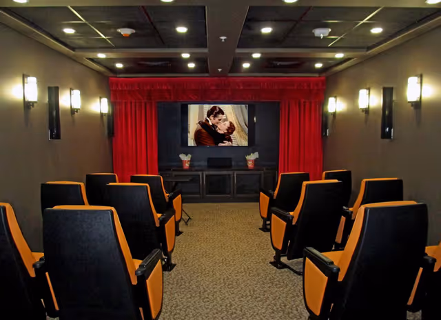 Small theater room with rows of black and orange cushioned seats facing a screen showing a romantic movie scene. The room has dark walls with wall-mounted lights and a red curtain framing the screen. Two containers of popcorn are placed on a cabinet below the screen.