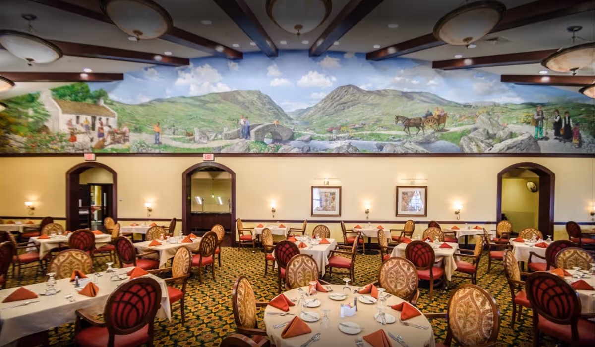 A spacious dining room with multiple round and rectangular tables covered with white tablecloths and set with plates, glasses, and folded orange napkins. The chairs have ornate upholstery in red and gold patterns. The walls are decorated with a large mural depicting a scenic countryside with hills, a stone bridge, people, and a horse-drawn carriage. The ceiling has wooden beams and large circular light fixtures.
