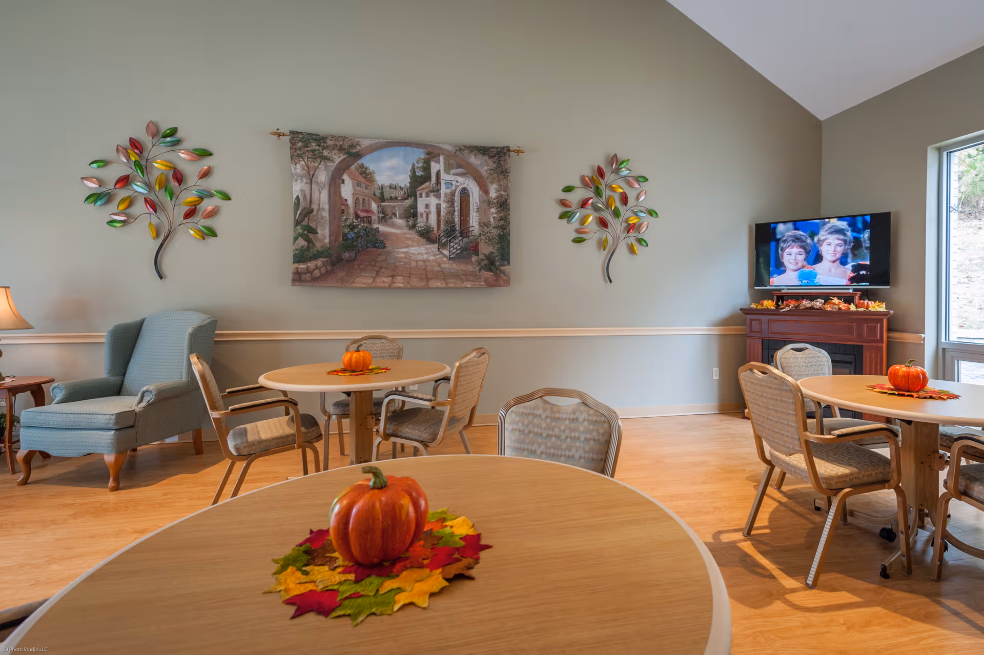 A cozy common area with round tables and chairs, each table decorated with a small pumpkin and colorful autumn leaves. There is a comfortable armchair with an ottoman on the left side, a wall tapestry depicting a scenic village street, and two decorative metal wall art pieces shaped like trees with colorful leaves. A flat-screen TV is mounted on the right wall above a wooden cabinet, and a large window lets in natural light.