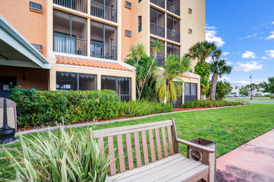 Outdoor view of a senior living facility with a beige bench in the foreground, green grass, palm trees, and shrubs along the building. The building has screened balconies and a tiled roof section under a clear blue sky.