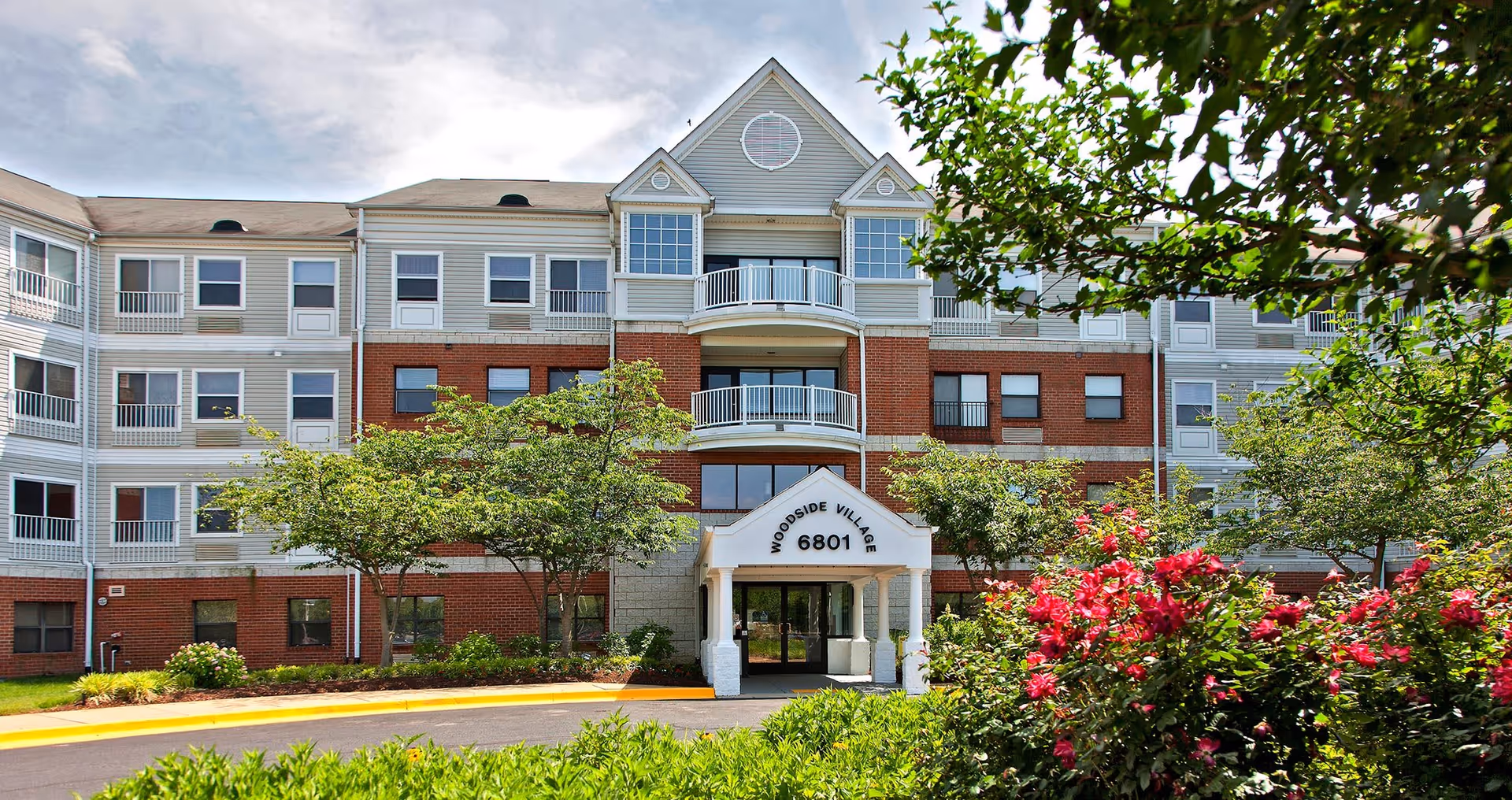 Front exterior of the Woodside Village multi-story senior living building with an entrance canopy, balconies, and landscaped shrubs.