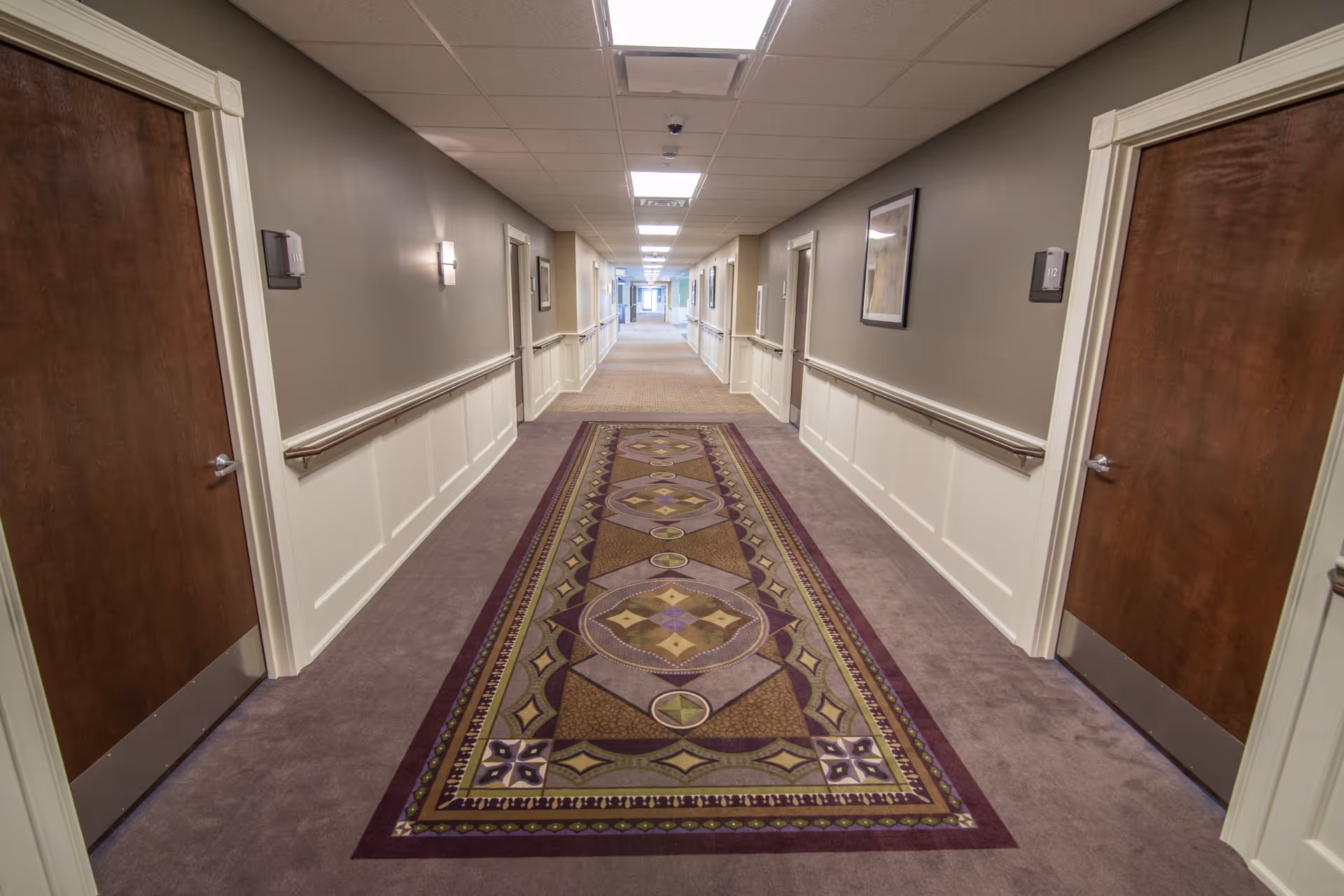 A long, well-lit hallway in a senior living facility with carpeted floors and a decorative runner rug in the center. The walls are painted gray with white wainscoting and handrails on both sides. Several wooden doors line the hallway, each with a room number plaque. Framed artwork is hung on the walls, and ceiling lights illuminate the corridor.