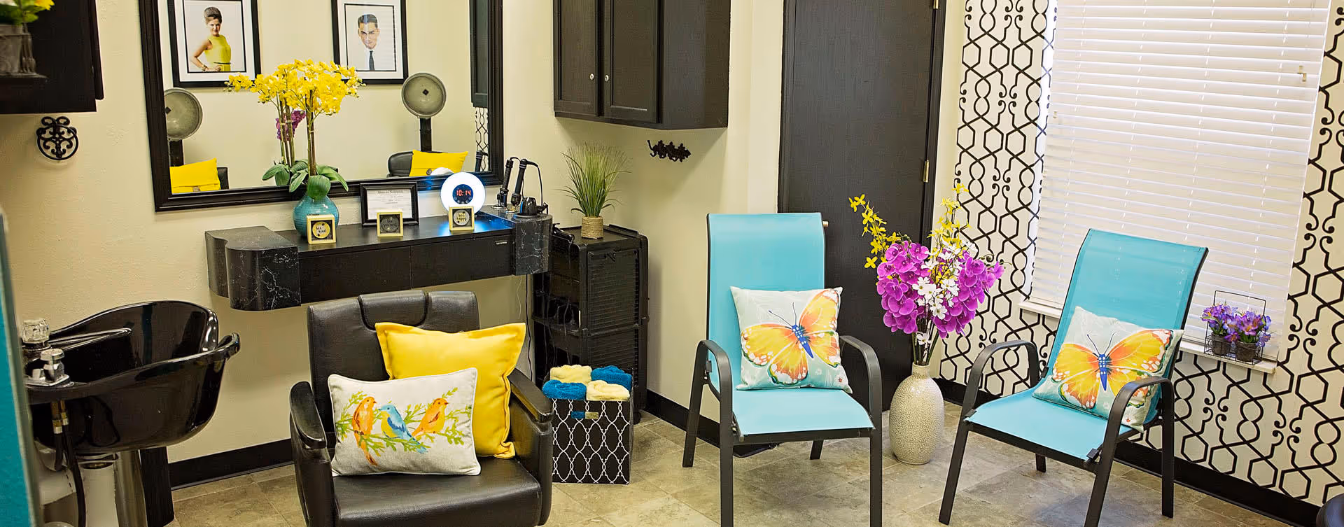 A brightly lit salon area with a black hair washing sink and a black salon chair with yellow and bird-themed pillows. Two turquoise chairs with butterfly-themed pillows are positioned near a window with patterned curtains. A vase with purple and yellow flowers sits between the chairs, and a large mirror hangs on the wall above a black floating shelf holding various small items and decorations.