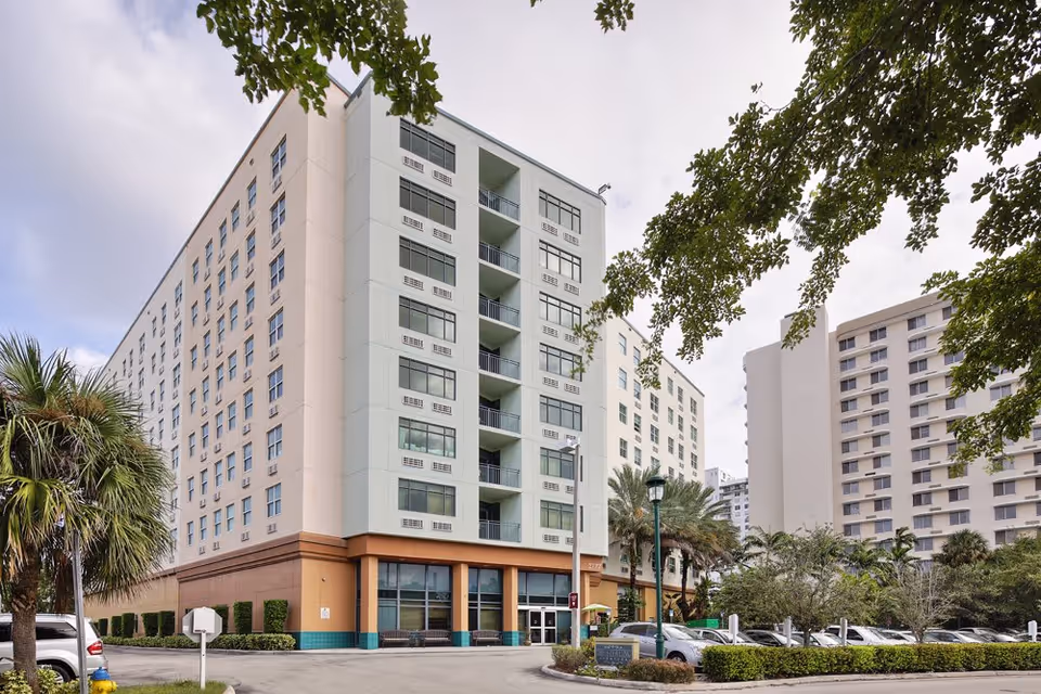 Exterior view of The Sterling Aventura, a multi-story senior living facility with large windows and balconies. The building is surrounded by palm trees and a parking lot with several cars. Other tall buildings are visible in the background under a partly cloudy sky.