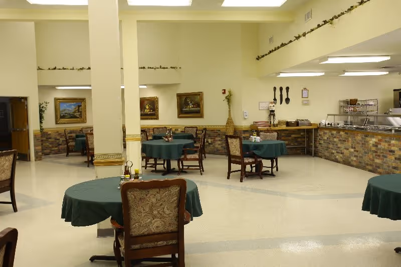 Dining room with round tables draped in green tablecloths, upholstered chairs, and a buffet counter along the right wall.