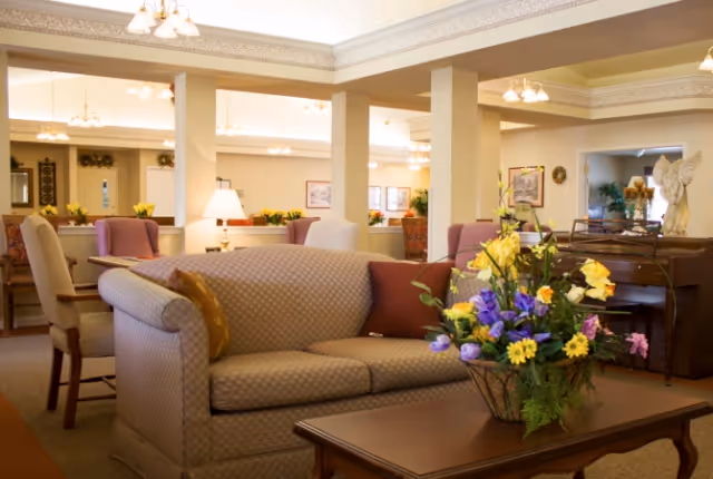 A cozy living room area in a senior living facility with a patterned beige sofa, a wooden coffee table with a colorful flower arrangement, several chairs, lamps, and decorative elements including framed pictures and a statue of an angel on a piano.