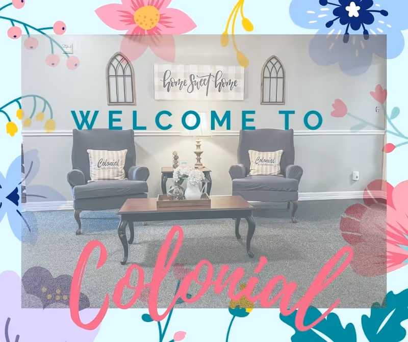 A cozy sitting area with two gray armchairs, each with a pillow that says 'Colonial Nursing & Rehabilitation'. Between the chairs is a small table with a lamp and decorative items. Above the chairs is a wall decoration with the phrase 'home sweet home' and two window-shaped wall hangings. The floor is carpeted and there is a wooden coffee table in front of the chairs. The image has floral graphics and the text 'WELCOME TO Colonial' overlaid.