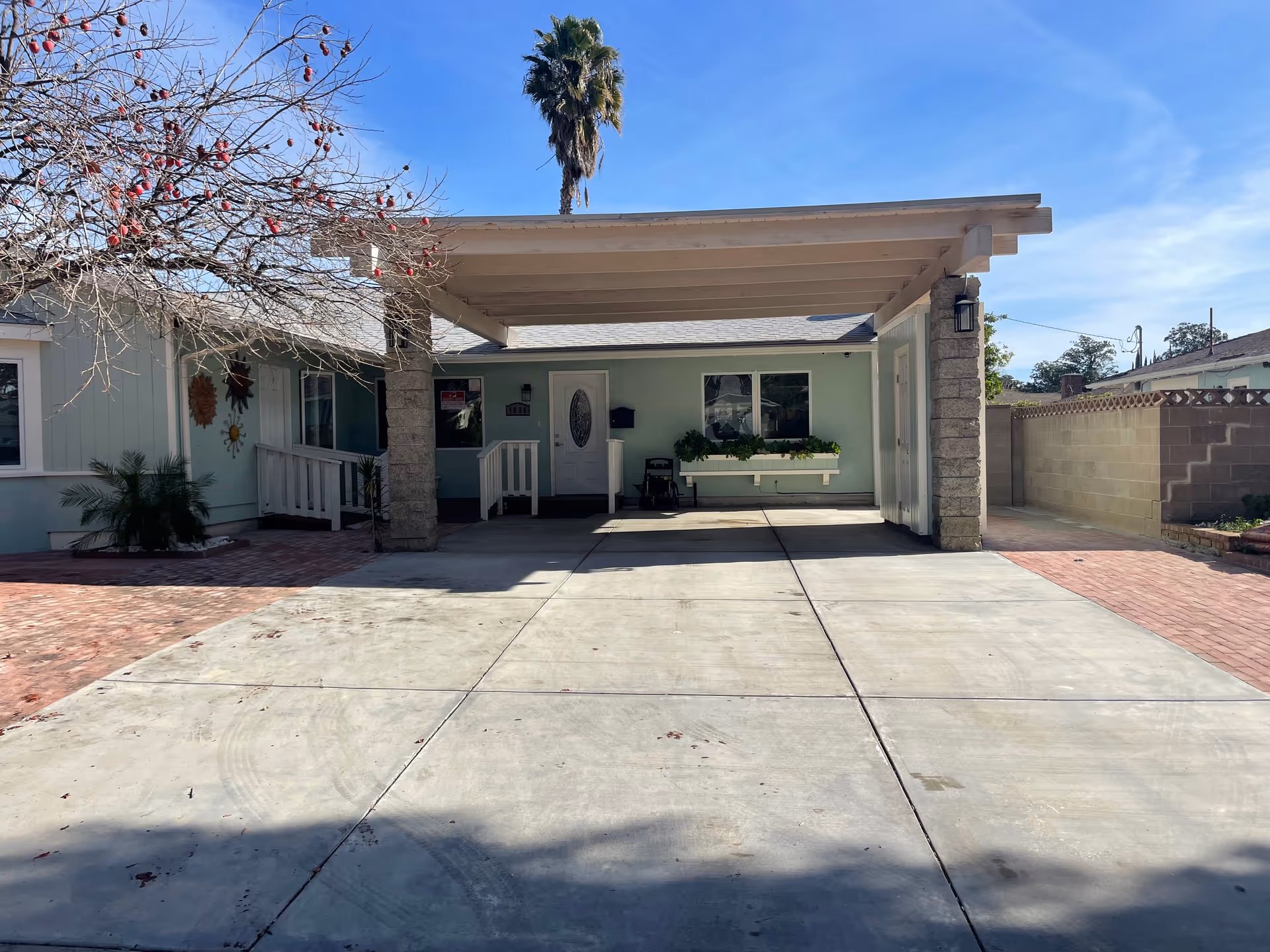 Front exterior view of a single-story assisted living facility with a covered carport supported by stone pillars. The building is light green with white trim, featuring a white front door with an oval glass window and a window box with plants. There is a palm tree visible behind the building and a leafless tree with red berries on the left side. The driveway is concrete with brick accents on the sides.