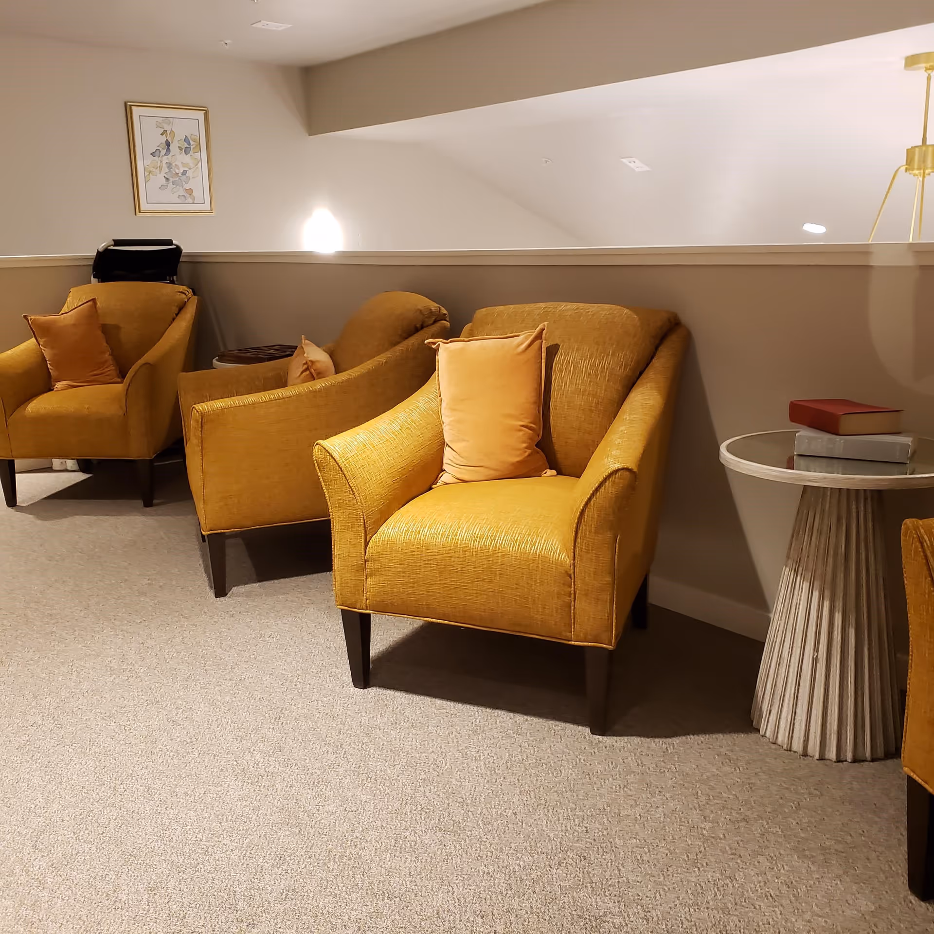 A cozy seating area with three mustard yellow armchairs, each with a matching pillow, arranged along a half wall. There is a round side table with a fluted base holding two books, and a framed floral artwork hangs on the wall above one of the chairs. The carpet is light beige and the lighting is warm.
