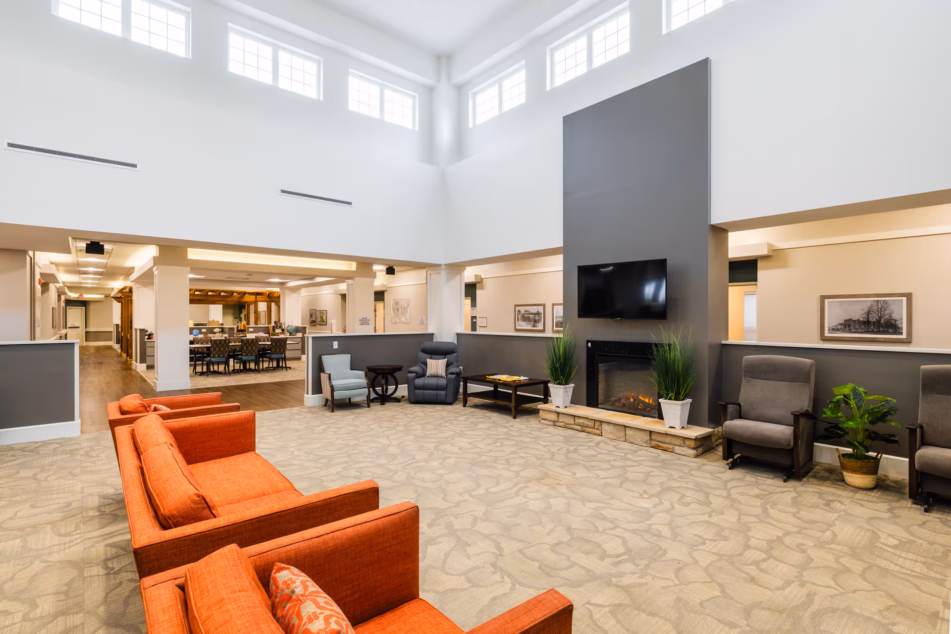 A spacious and well-lit living room area in a senior living facility with high ceilings and multiple windows near the ceiling. The room features orange sofas, gray armchairs, a fireplace with a TV mounted above it, and several potted plants. In the background, there is a dining area with tables and chairs.