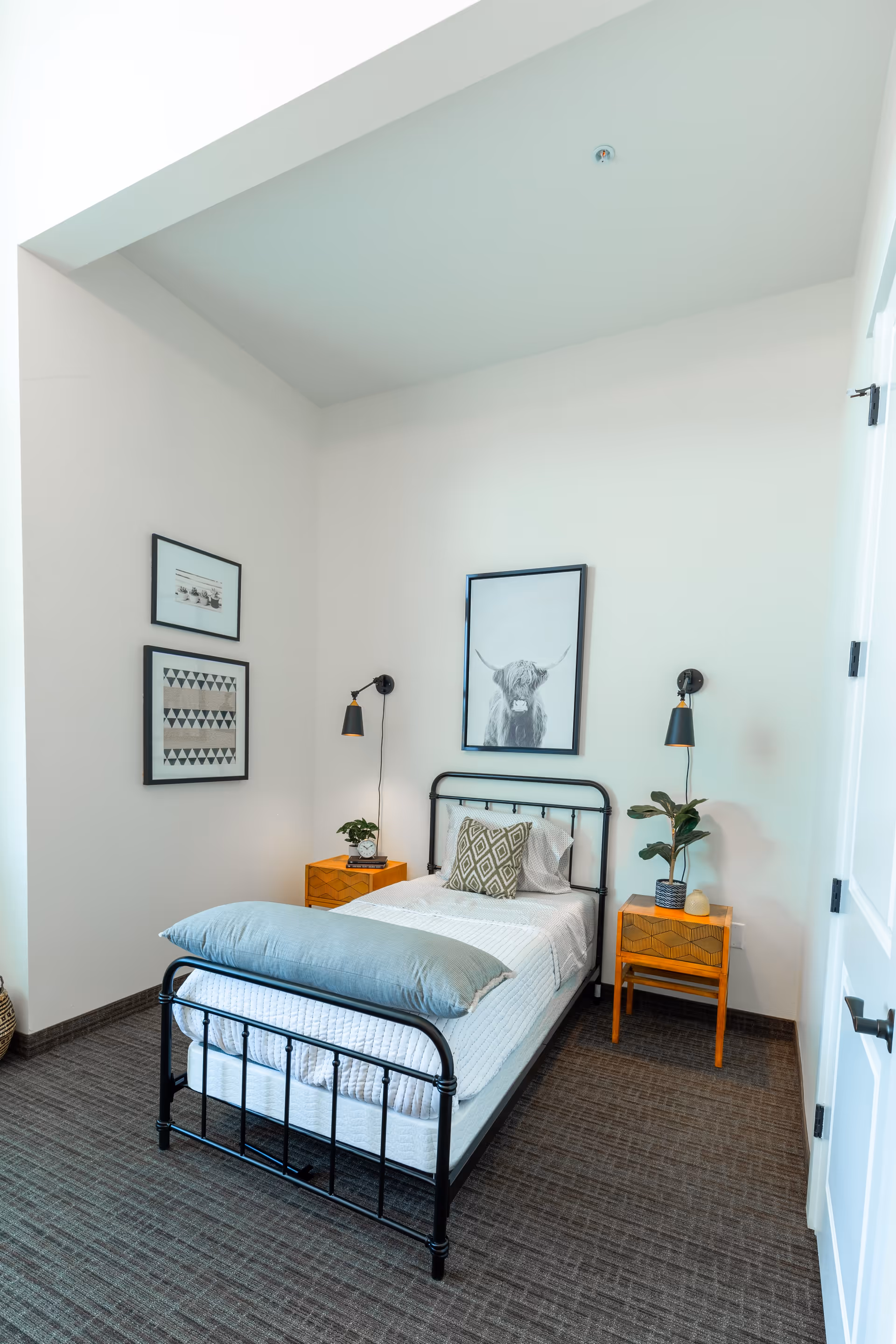 A neatly arranged bedroom with a single black metal bed frame, white bedding, a gray pillow, and a decorative cushion. There are two wooden nightstands on either side of the bed, each with a small plant and a wall-mounted black lamp above. The walls are white with three framed pictures, including one of a highland cow above the bed. The floor is carpeted in a dark pattern.