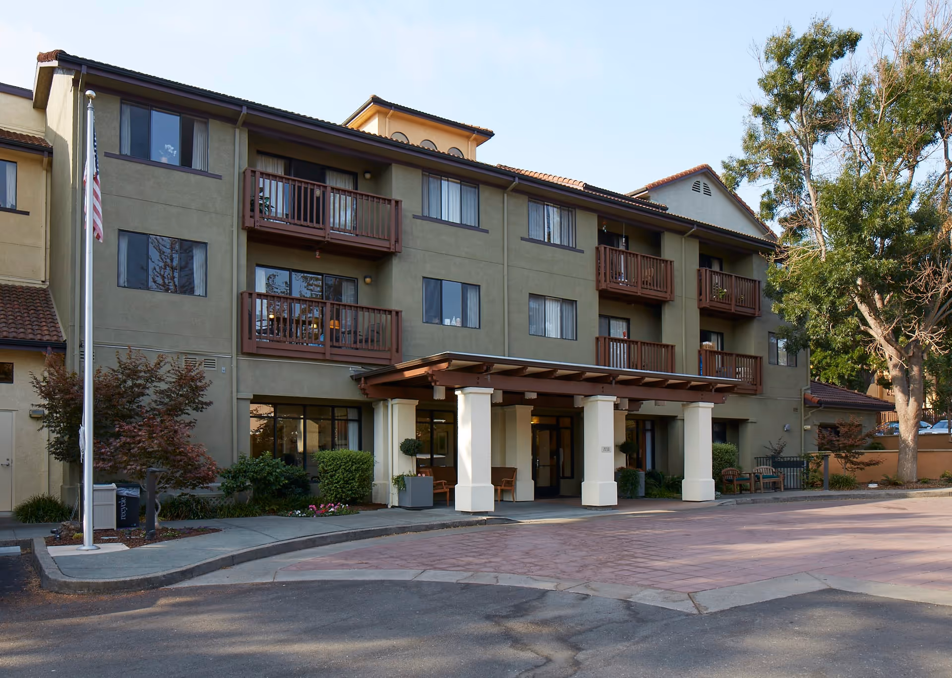 Exterior view of a three-story senior living facility building with balconies, an entrance canopy supported by white columns, landscaping with trees and bushes, and an American flag on a flagpole.