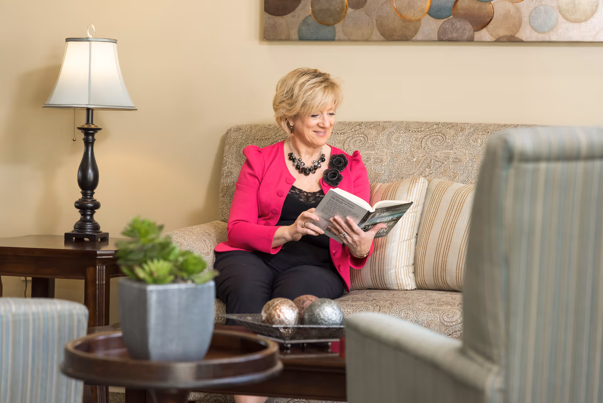 A woman in a pink jacket sits on a patterned sofa reading a book in a cozy living room with a lamp and coffee table.