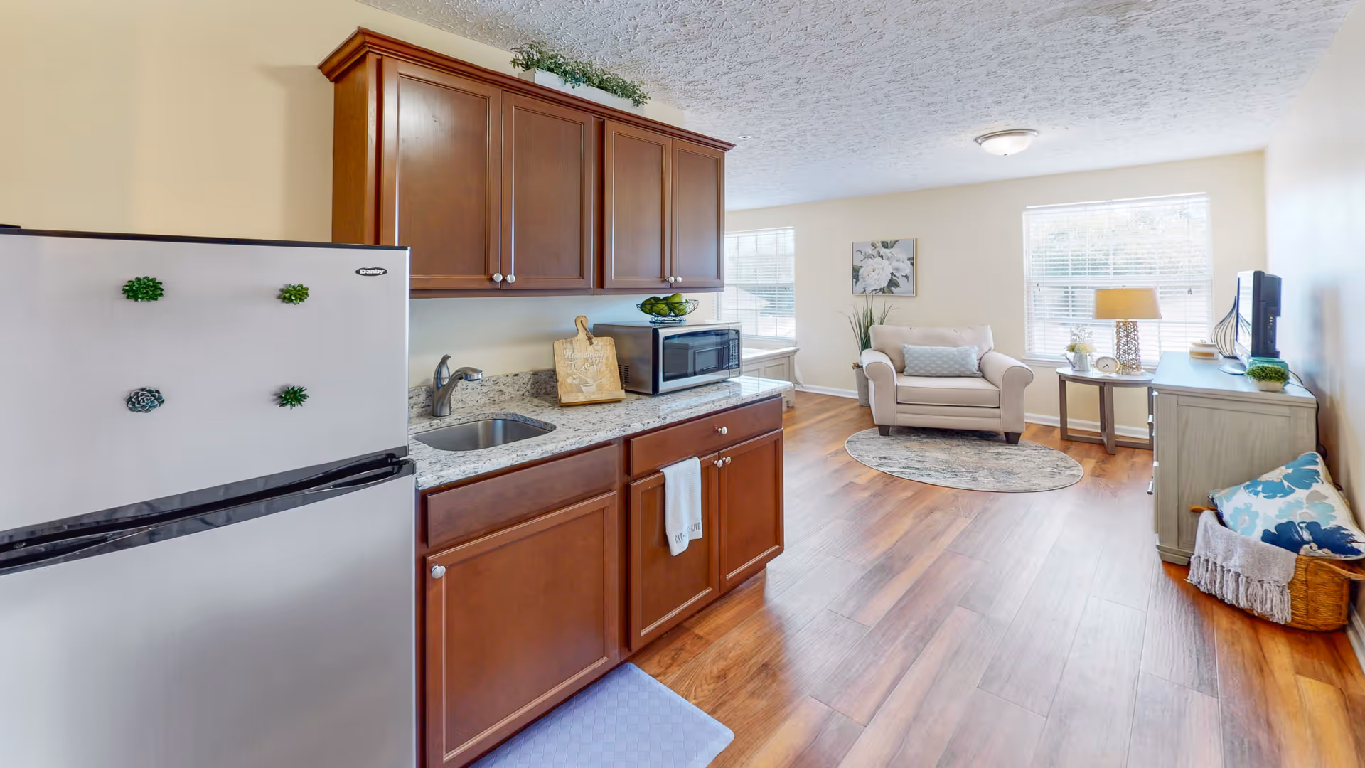Open-plan room with a small kitchenette on the left and a sitting area with an armchair, side table, and TV on the right.