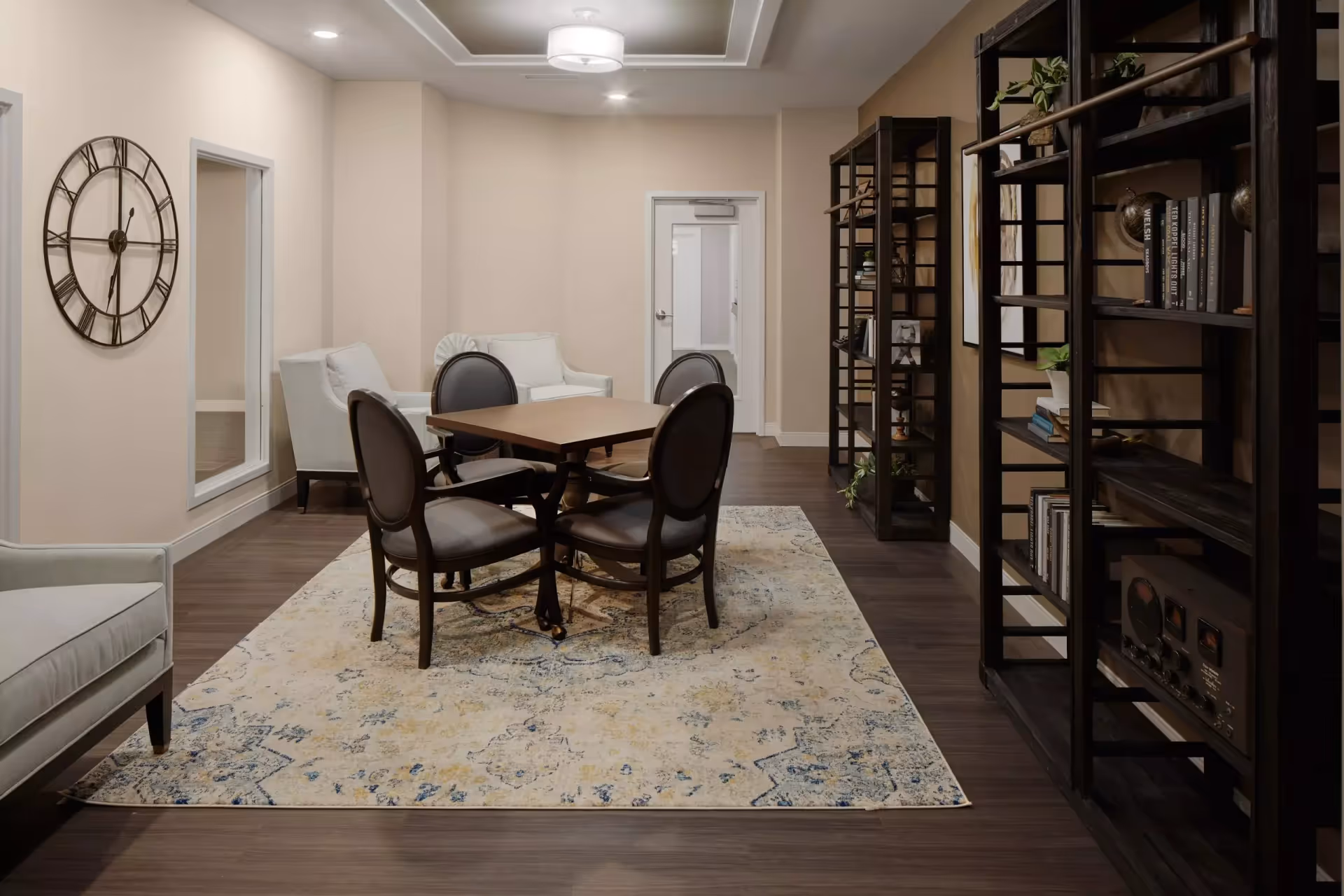 A cozy interior room with a square wooden table surrounded by four cushioned chairs on a patterned area rug. There are two armchairs in the background near a wall with a large round clock and a tall mirror. On the right side, there are dark wooden open shelves with books and decorative items. The room has soft lighting and a neutral color palette.