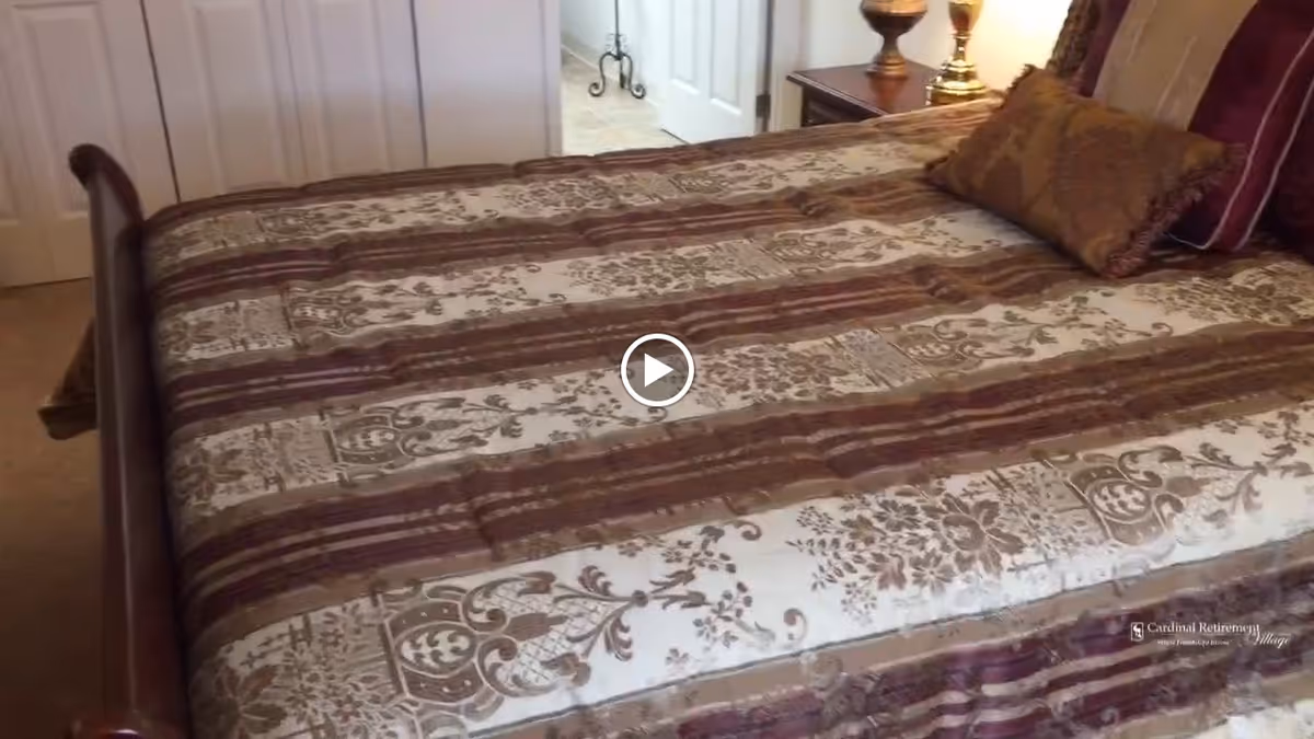 A bedroom featuring a bed with a patterned bedspread in shades of brown and cream, two decorative pillows, a wooden headboard, a bedside table with a lamp, and a partially open door leading to another room.