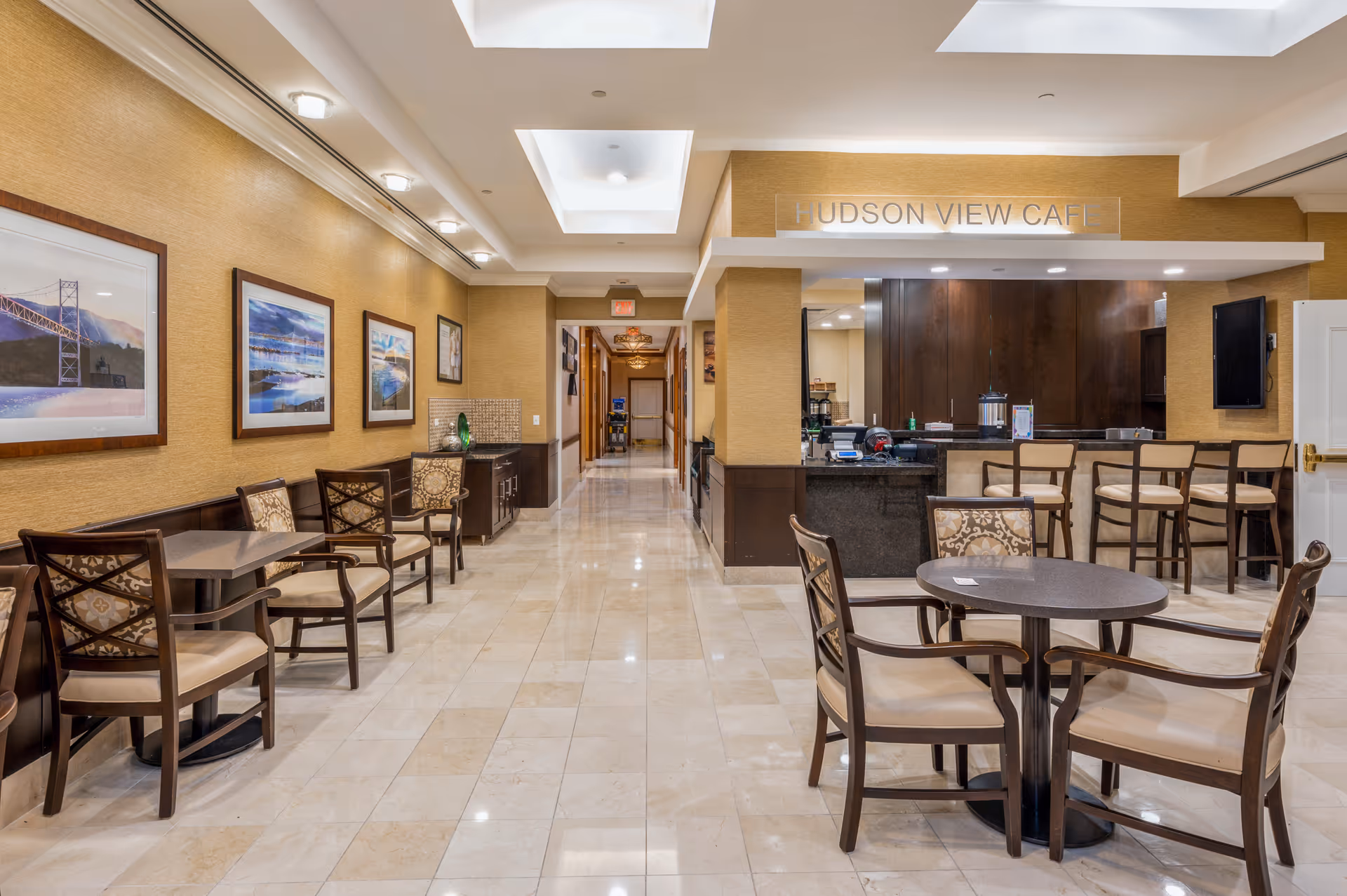 Interior view of a dining area named Hudson View Café with several tables and chairs arranged neatly. The walls are decorated with framed pictures, and the floor is tiled with light-colored marble. There is a counter with bar stools on the right side and a hallway leading further into the building.
