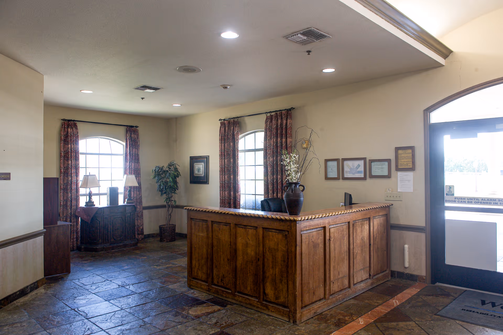 Reception area with a wooden front desk, two large windows with patterned curtains, a potted plant, framed pictures on the wall, and a glass door letting in natural light.