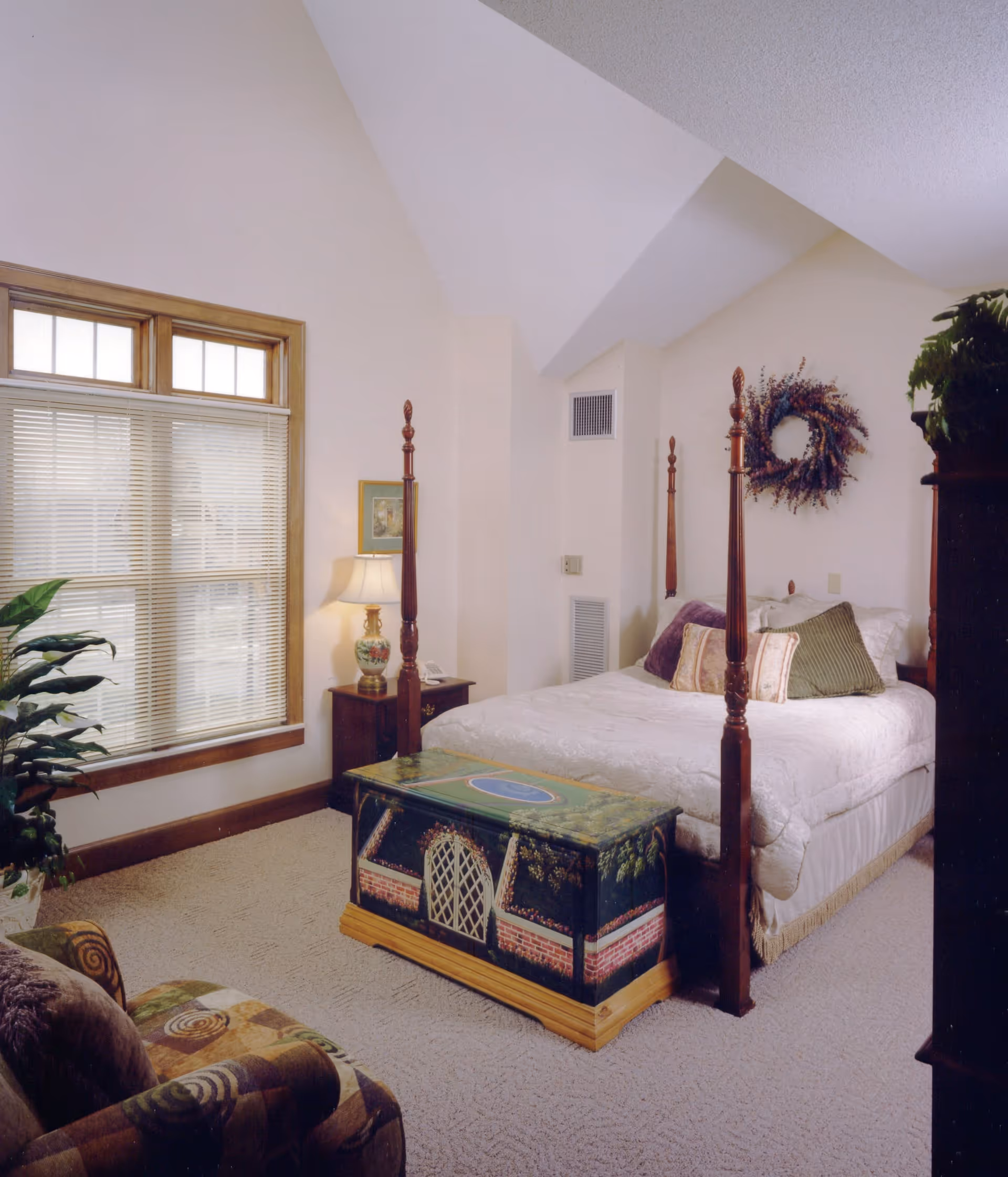 Bright bedroom with a four-poster bed, decorative chest at the foot, bedside table with lamp, window with blinds, and an upholstered chair.