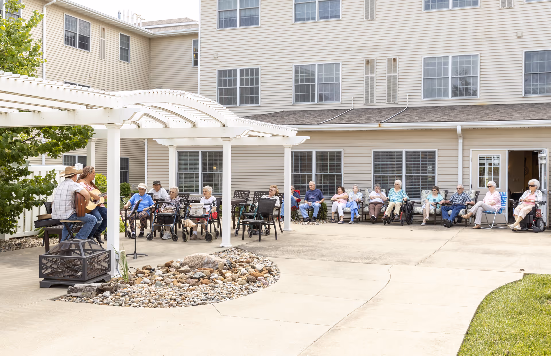 A group of elderly people sitting outdoors in a semi-circle on chairs and wheelchairs, watching two musicians playing guitars under a white pergola. The setting is a courtyard of a beige multi-story building with large windows and a concrete pathway surrounding a small rock garden.