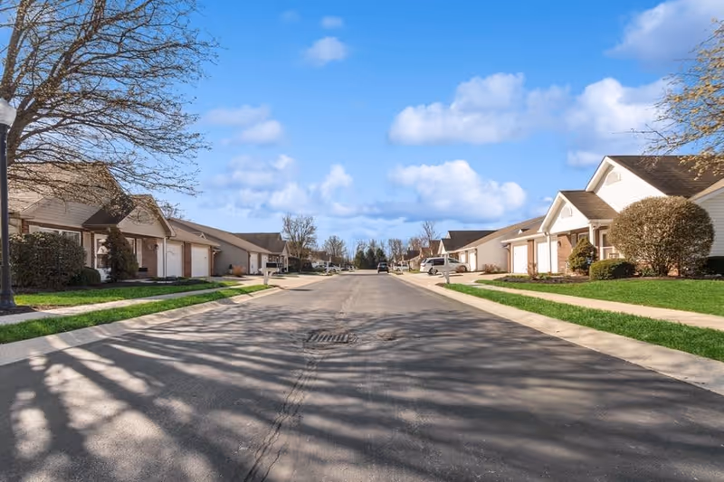 A residential street lined with single-story houses featuring garages, manicured lawns, and some trees under a blue sky with scattered clouds.