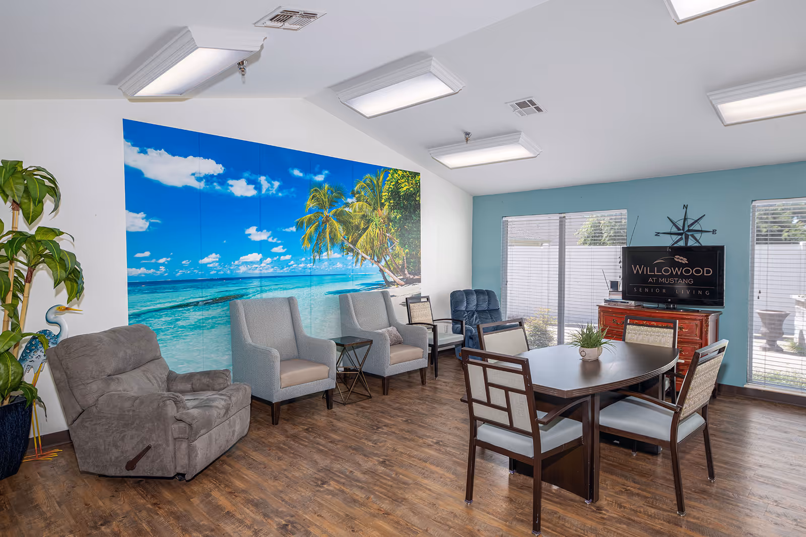 Bright senior living common room with chairs, a dining table, a tropical beach wall mural, and a TV console.