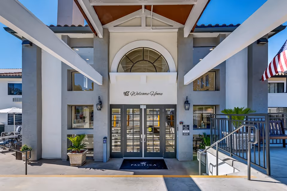 Entrance of a senior living facility with double glass doors under an archway. Above the doors is a sign that reads 'Welcome Home.' There are potted plants on either side of the entrance, outdoor seating visible to the left, and an American flag on the right side. The building exterior is light gray with white accents.