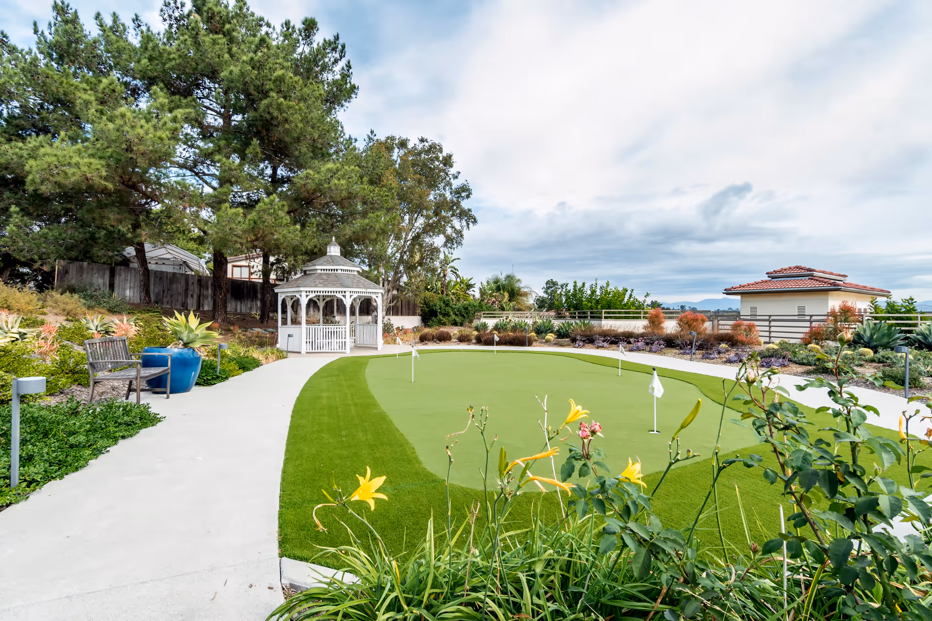 Outdoor garden area with a putting green, a white gazebo, a wooden bench, and various plants and flowers under a partly cloudy sky.