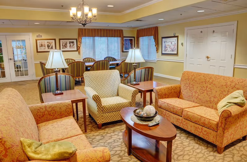 Cozy senior living common room with sofas, an armchair, coffee table, lamps, and a dining table in the background.