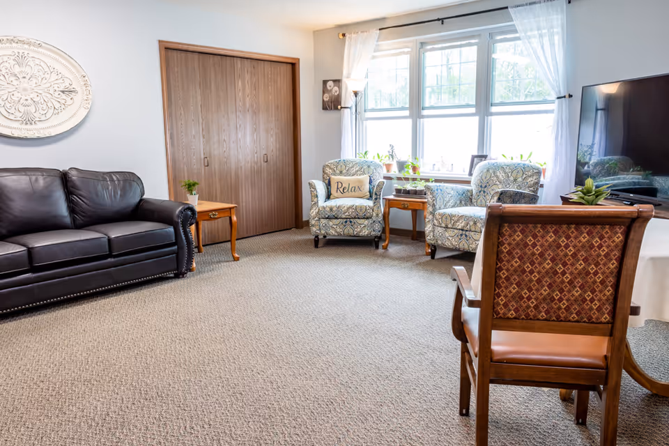 A cozy living room with a dark leather sofa on the left, two patterned armchairs with a 'Relax' pillow near a large window with white curtains, a wooden side table with a plant, a wooden chair with a patterned backrest in the foreground, and a flat-screen TV on the right.