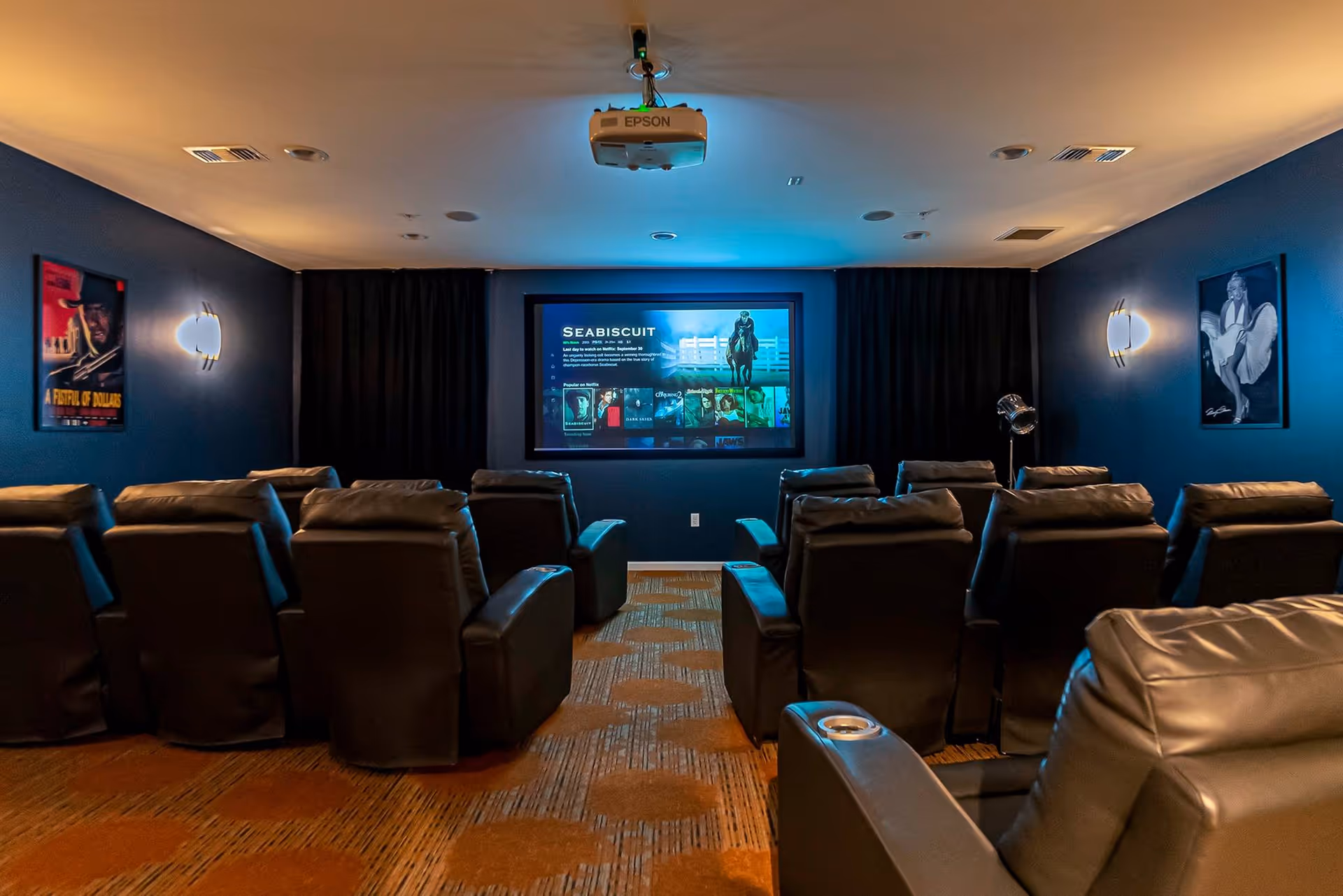 A cozy movie theater room with multiple rows of black leather recliner chairs facing a large screen displaying the movie selection menu. The walls are painted dark blue and decorated with framed movie posters. There are wall-mounted lights on both sides of the room and a ceiling-mounted Epson projector.